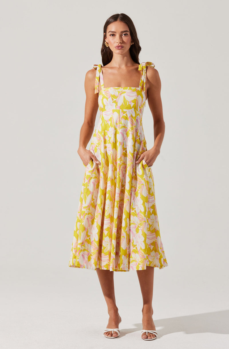 A woman is wearing a sleeveless, ankle-length dress with a fitted bodice and a flared skirt. The dress features shoulder straps tied into bows on each shoulder and a vibrant floral pattern with large pink and white flowers and green leaves on a yellow background. She is also wearing white strappy high-heeled sandals and gold hoop earrings.