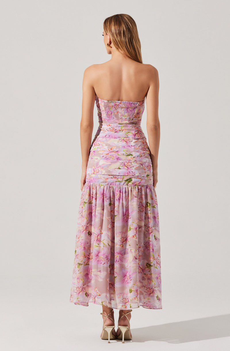 A woman is wearing a strapless, backless dress with a fitted bodice and a tiered, flowing skirt. The dress features a pink floral pattern with shades of purple, green, and white, creating a soft, romantic look. She is also wearing nude-colored high heels.