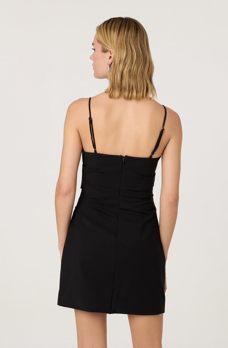 A woman is wearing a sleeveless black dress with thin adjustable spaghetti straps. The dress is fitted at the bodice and has a visible zipper running down the back. The hemline of the dress falls above the knees.