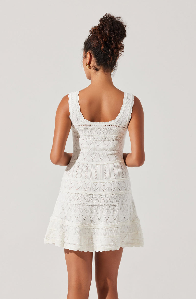 A woman is wearing a sleeveless, white dress with a fitted waist and a flared skirt. The dress features intricate lace and embroidered patterns throughout, with a scalloped hemline and wide, scalloped shoulder straps.