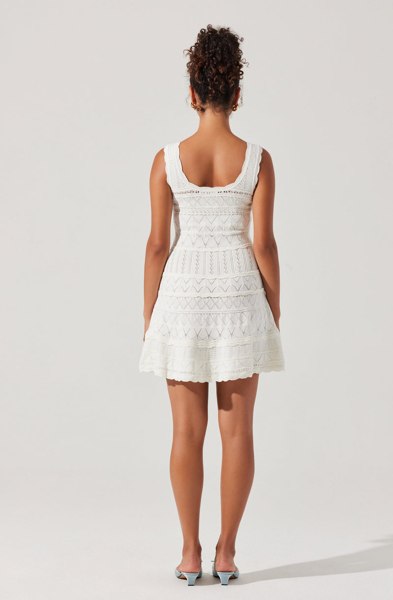 A woman is wearing a sleeveless, knee-length white dress with lace and embroidered patterns. The dress has wide straps and a scoop neckline at the back. She is also wearing light-colored high-heeled shoes.