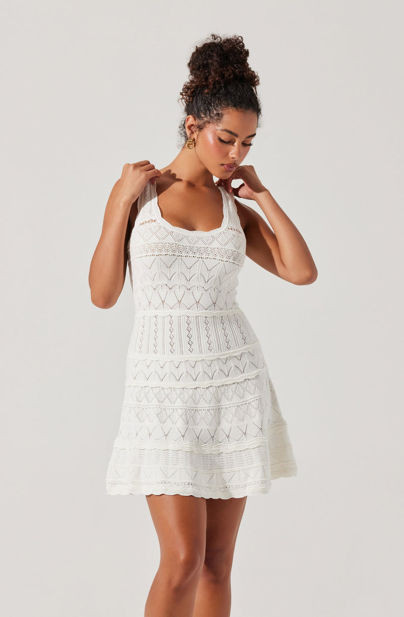 A woman is wearing a sleeveless, knee-length white dress with intricate lace and embroidery patterns. The dress has a fitted bodice with a scoop neckline and layered sections with different lace designs, creating a textured appearance. She has her hair styled in a curly updo and is holding the straps of her dress with both hands, looking down with a contemplative expression.