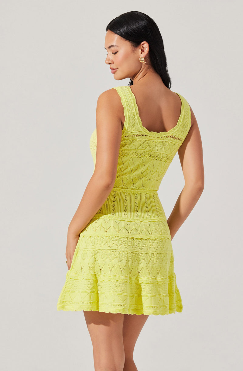A woman is wearing a sleeveless, bright yellow dress with intricate geometric lace patterns throughout. The dress has wide straps, a fitted bodice, and a flared skirt that reaches above the knees. The fabric appears lightweight and textured with detailed embroidery.