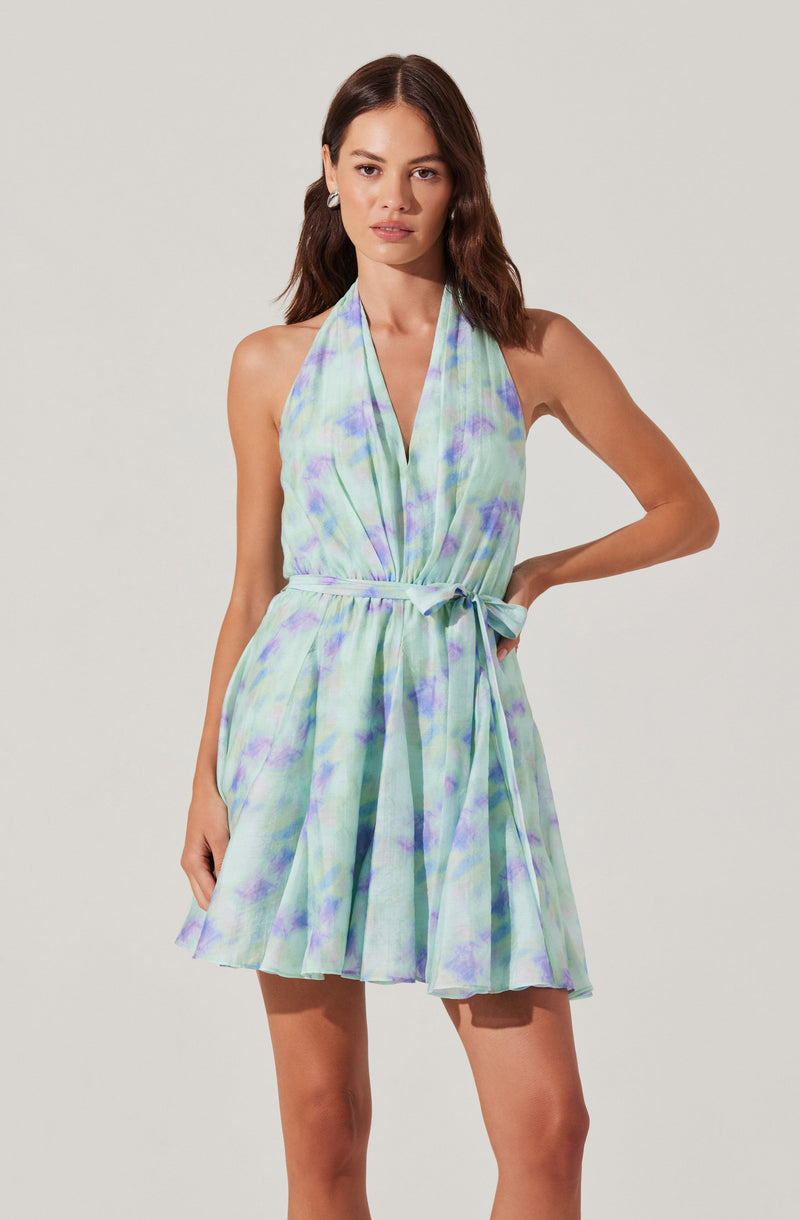 A woman is wearing a sleeveless, halter-neck dress with a waist tie. The dress has a light, flowing fabric with a pastel watercolor pattern in shades of blue, green, and purple.
