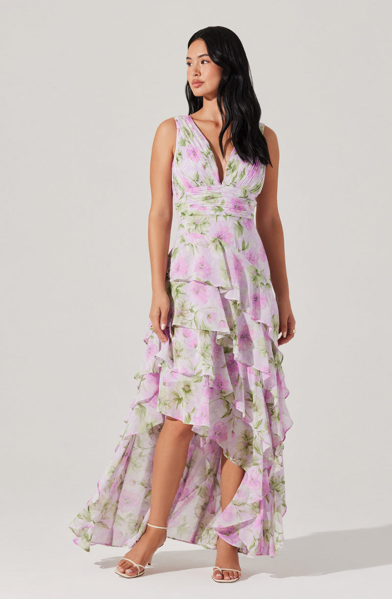 A woman is wearing a sleeveless, V-neck maxi dress with a fitted waist. The dress features a light-colored floral pattern with shades of pink and green, layered with ruffled tiers down the length of the skirt. She is wearing open-toed, strappy high heels.