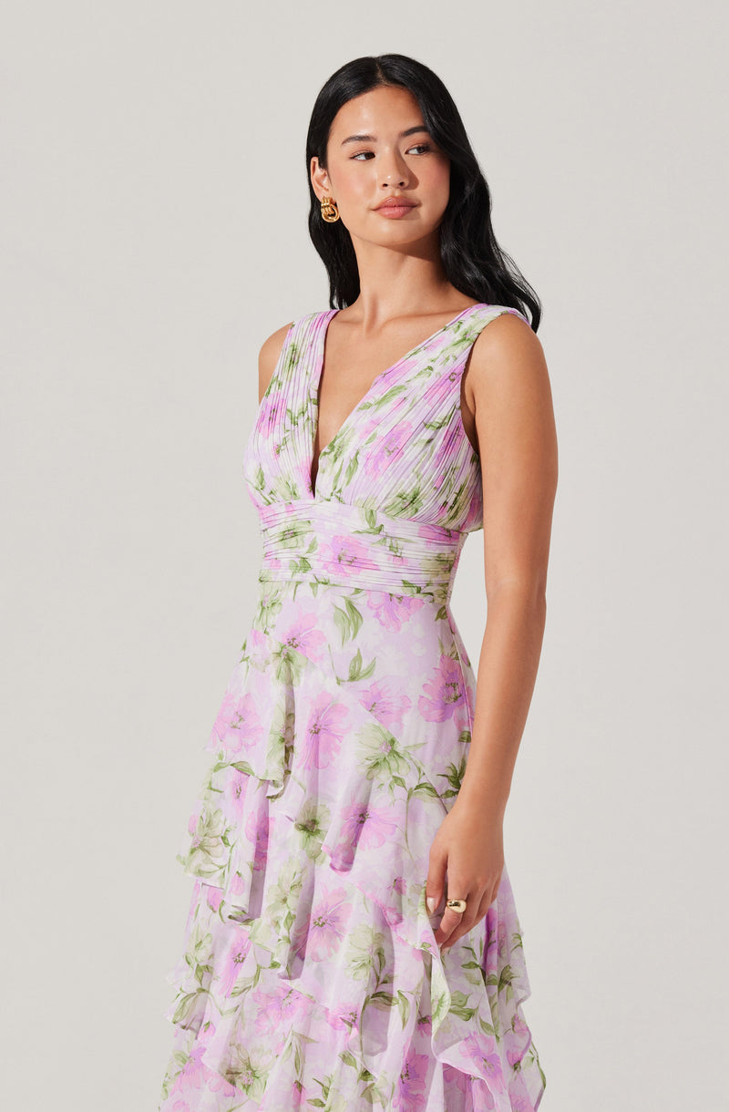 A woman is wearing a sleeveless, V-neck dress with a fitted bodice that features a gathered design and a waistline accentuated with a wide band. The dress has a floral pattern with pink and green flowers on a white background. The skirt is tiered with ruffled layers, creating a flowing and layered appearance.