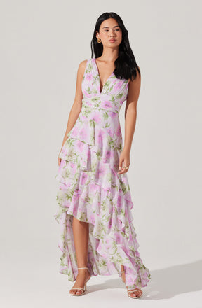 A woman is wearing a sleeveless, V-neck dress with a fitted bodice and a flowy, tiered skirt. The dress features a pastel floral pattern in shades of pink, green, and white. She is also wearing high-heeled sandals and gold hoop earrings.