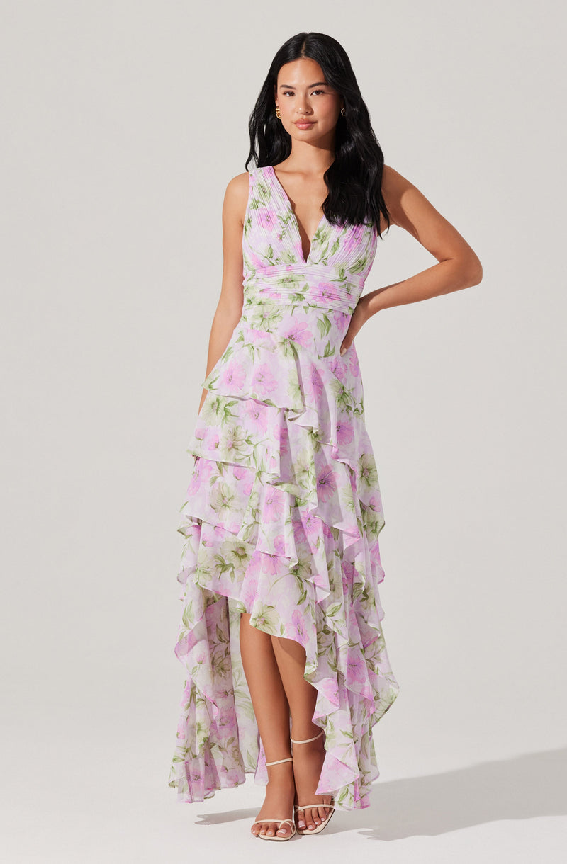 A woman is wearing a sleeveless, V-neck, floral-print dress with shades of pink and green on a white background. The dress features a fitted bodice and layered ruffles cascading down the length, creating a high-low hemline with the front ending above the ankles and the back flowing to the floor. She is also wearing strappy high-heeled sandals.