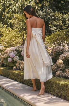 A woman is standing barefoot on a stone edge next to a swimming pool, wearing a strapless white dress with a bow tied at the back. The dress is long and flowing, reaching mid-calf, and appears to be made of a light, silky fabric. She has long, dark hair that falls past her shoulders. The background features lush greenery and blooming hydrangea flowers in shades of pink and white.
