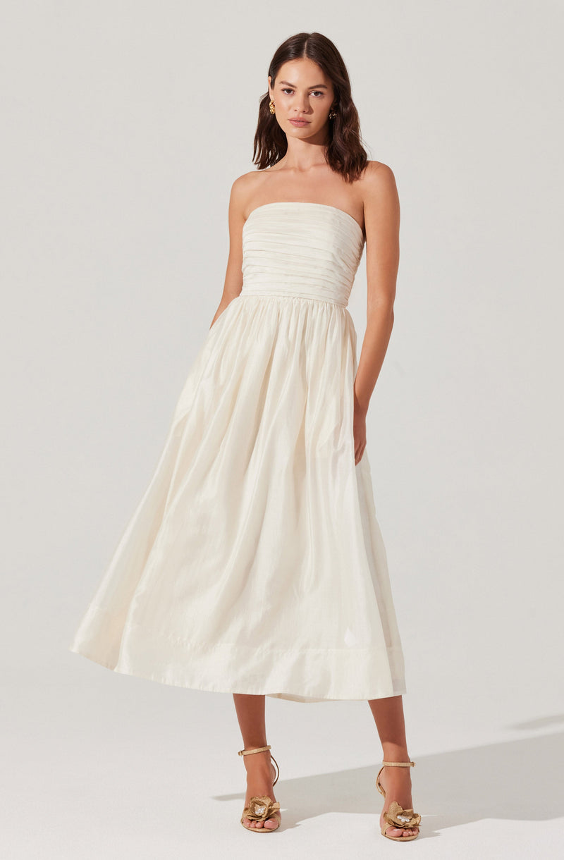 A woman is wearing a strapless, off-white, midi-length dress with a ruched bodice and a full, gathered skirt. She has on open-toed, high-heeled shoes with ankle straps and decorative bows.