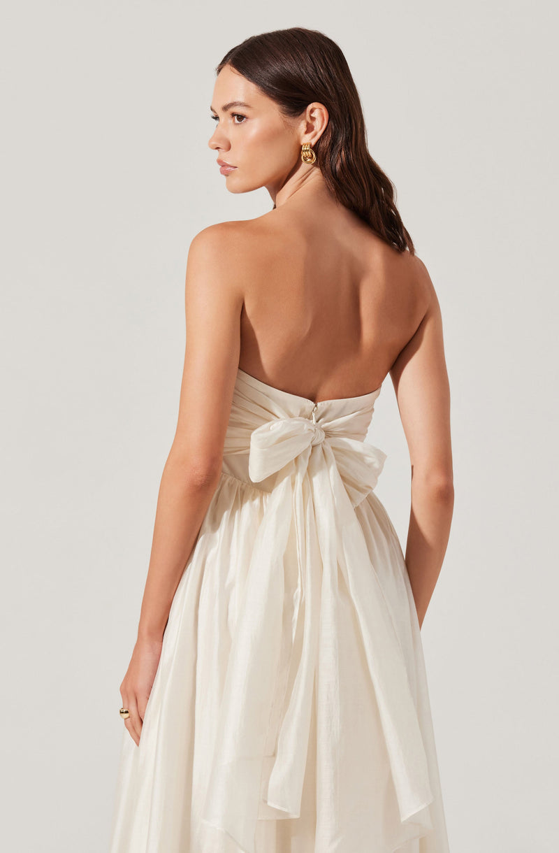A woman is wearing a strapless, off-white, satin dress with a large bow tied at the back. The dress has a gathered bodice and a flowing skirt. She has shoulder-length dark hair, gold earrings, and a ring on her left hand.