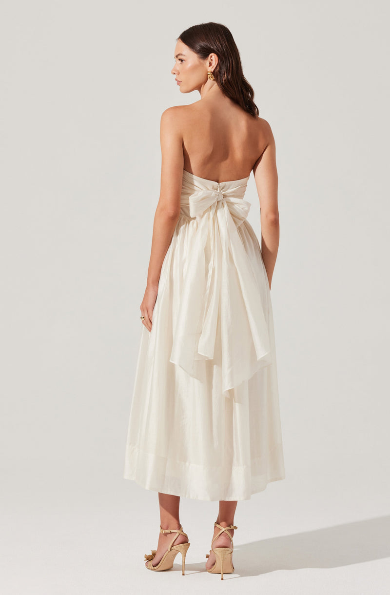 A woman is wearing a strapless, flowing, cream-colored dress with a large bow tied at the back. The dress has a fitted bodice and a full skirt that reaches mid-calf. She is also wearing beige high-heeled shoes with ankle straps.