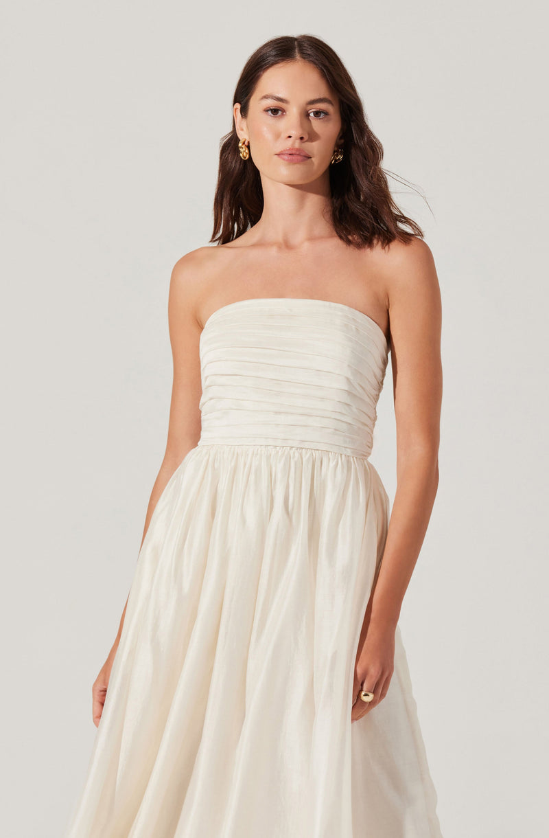 A woman is wearing a strapless, floor-length dress with a fitted, pleated bodice and a flowing skirt made of shiny, cream-colored fabric. She has dark, wavy hair and gold hoop earrings.
