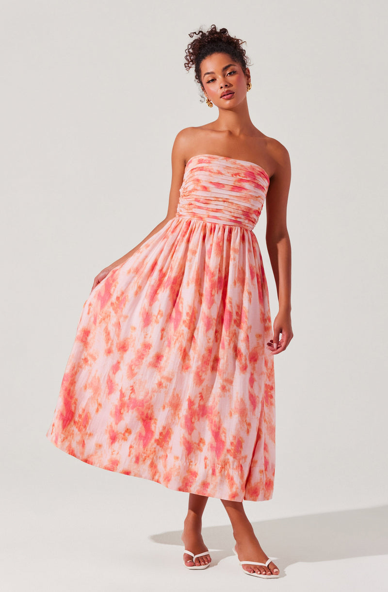 A woman is wearing a strapless, full-length dress with a fitted, ruched bodice and a flowing skirt. The dress features a pink and orange watercolor-like abstract pattern. She is also wearing white high-heeled sandals.
