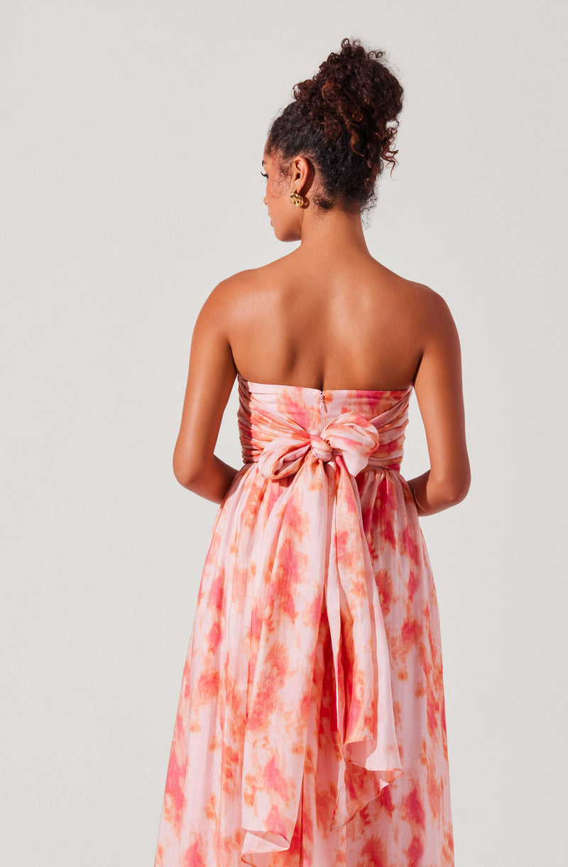 A woman is wearing a strapless, flowing dress with a watercolor-like pattern in shades of pink, orange, and white. The dress features a large bow at the back, tied at the waist, with fabric draping down. She has her hair styled in a high, curly bun and is accessorized with gold hoop earrings.