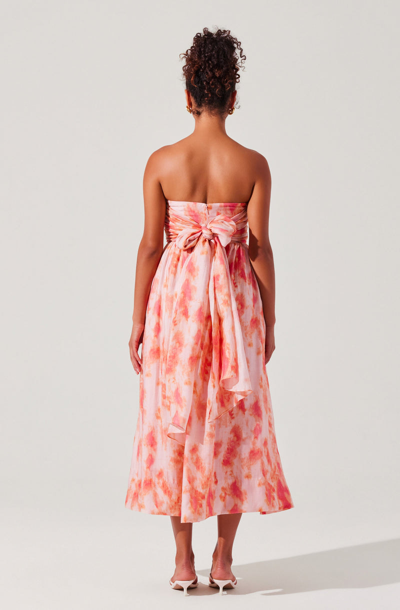 A woman is wearing a strapless, floor-length dress with a watercolor-like pattern in shades of pink and orange. The dress features a large bow tied at the back around the waist, with the fabric cascading down. She is also wearing a pair of light-colored high-heeled shoes.