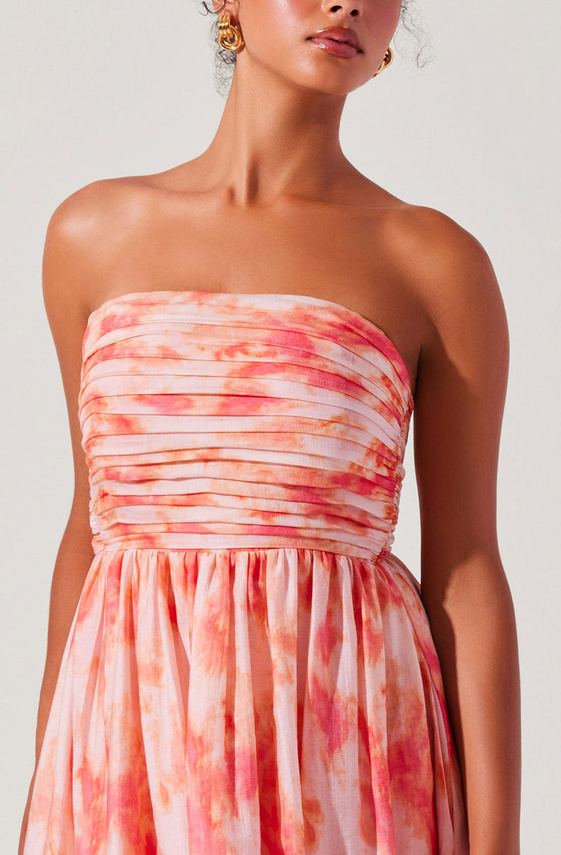 The dress is strapless with a fitted bodice featuring horizontal pleats. It has a flowing skirt with a pink and orange watercolor-like tie-dye pattern. The material appears lightweight and soft, giving a breezy, summery feel.