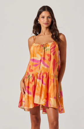 A woman is wearing a sleeveless, knee-length dress with spaghetti straps. The dress features a loose, flowy fit and has a vibrant, abstract pattern in shades of orange, pink, red, green, and beige.