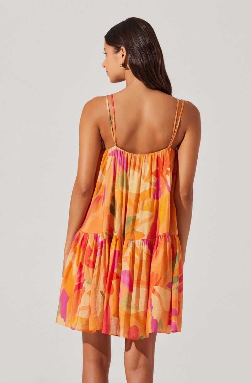 A woman is wearing a sleeveless dress with thin, adjustable spaghetti straps. The dress features a loose, flowing fit with a tiered design and a vibrant, abstract floral pattern in shades of orange, pink, yellow, and green.