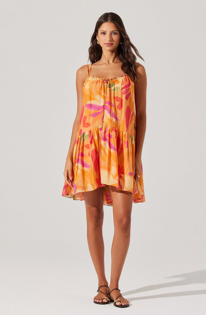 The dress is a sleeveless, above-the-knee style with thin adjustable spaghetti straps. It features a vibrant, multicolored abstract pattern predominantly in shades of orange, pink, and green. The dress has a loose, flowy fit with a ruffled hemline, creating a casual and summery look.