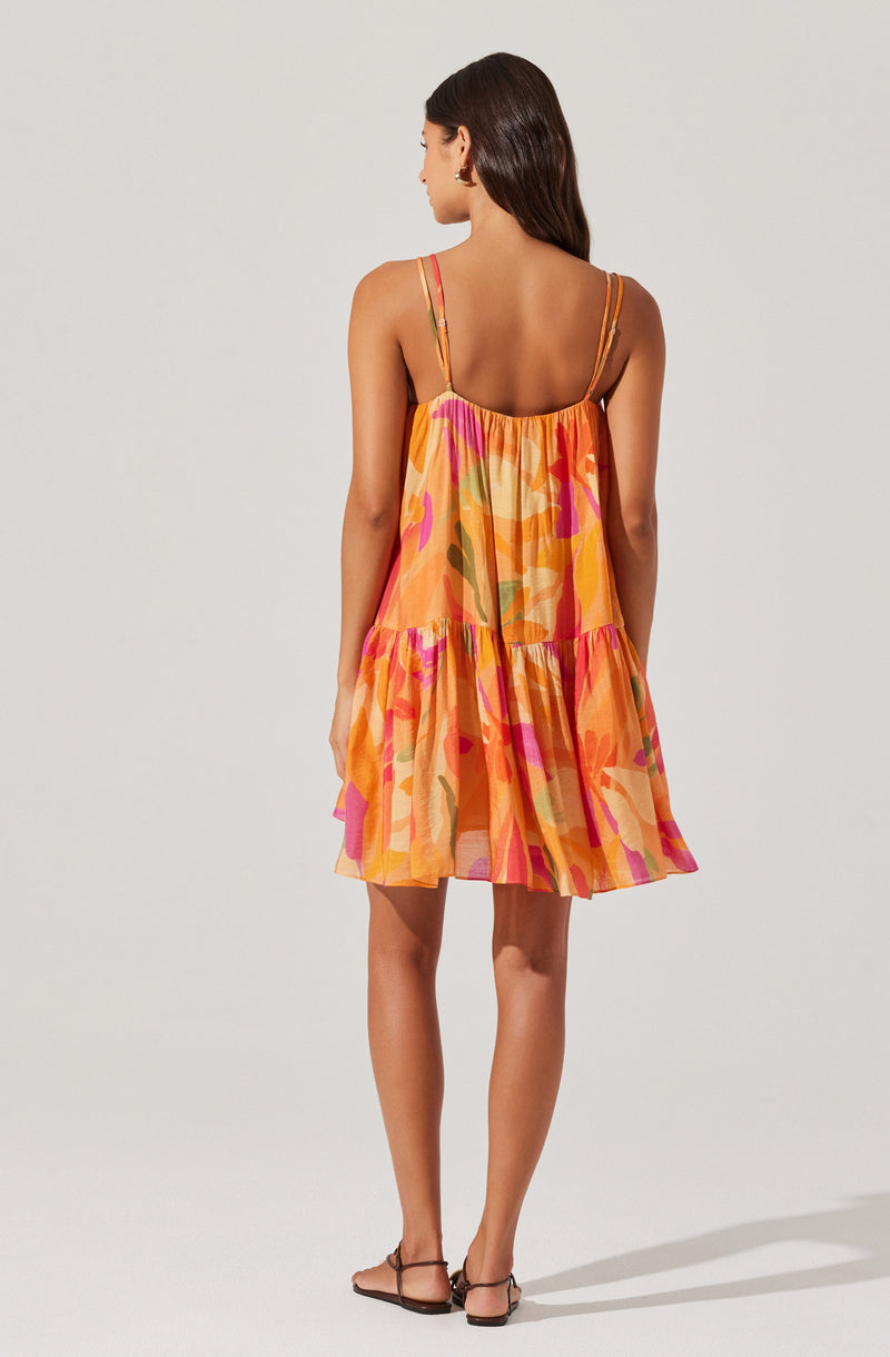 A woman is wearing a loose-fitting, sleeveless sundress with spaghetti straps. The dress features a colorful abstract pattern with shades of orange, pink, yellow, and green, and has a tiered, ruffled hem.