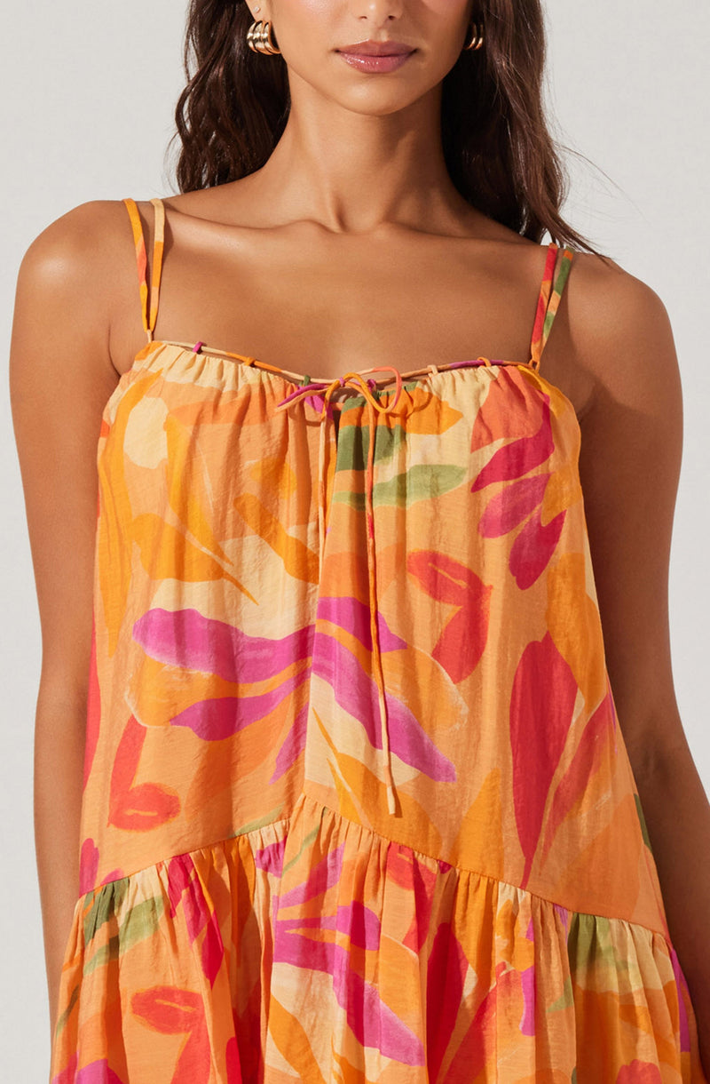 A woman is wearing a sleeveless dress with thin spaghetti straps. The dress features a vibrant, abstract floral pattern in shades of orange, pink, red, green, and beige. The fabric appears lightweight and flowy, with a gathered ruffle detail near the waist and a tied bow at the neckline.
