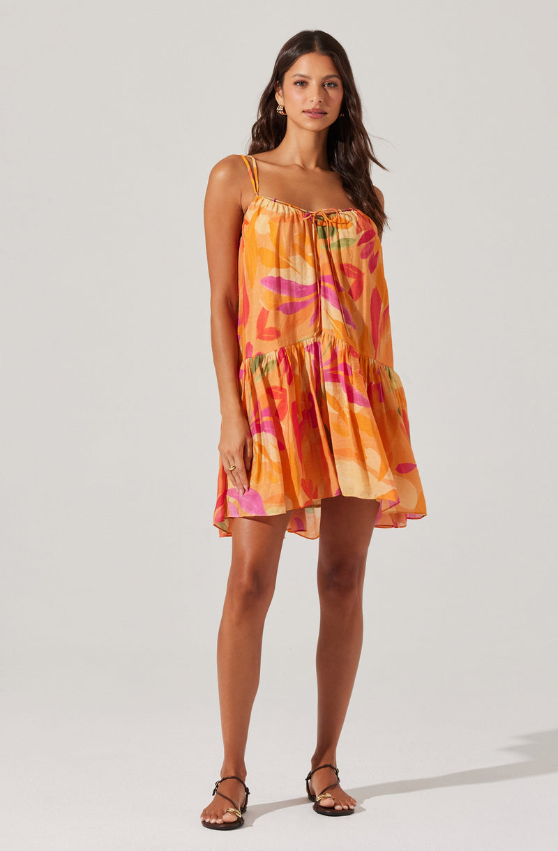 A woman is wearing a sleeveless, knee-length sundress with thin shoulder straps. The dress features an elasticized neckline with a drawstring tie and has a loose, flowy fit. The fabric has a colorful, abstract pattern with shades of orange, pink, yellow, and green. She is also wearing flat, strappy sandals.