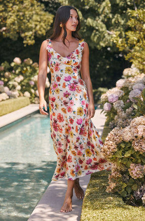A woman is wearing a sleeveless, ankle-length dress with a floral pattern featuring pink, yellow, orange, and purple flowers on a white background. The dress has a ruffled neckline and a flowy, lightweight fabric.