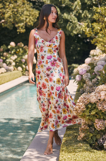 A woman is wearing a sleeveless, ankle-length dress with a floral pattern featuring pink, yellow, orange, and purple flowers on a white background. The dress has a ruffled neckline and a flowy, lightweight fabric.