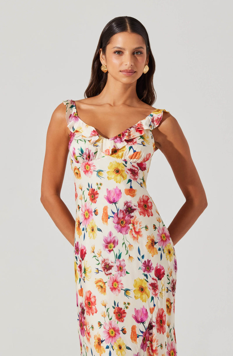 A woman is wearing a sleeveless, floral dress with ruffled shoulder straps. The dress features a colorful pattern of pink, yellow, orange, and purple flowers with green leaves on a light background. She accessorizes with large, gold, circular earrings and has long, dark, wavy hair.