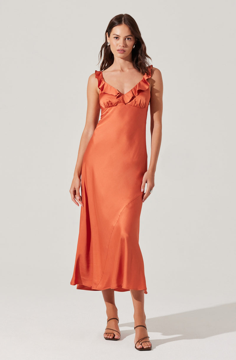 A woman is wearing a long, satin dress in a warm, burnt orange color. The dress features ruffled shoulder straps and a V-shaped neckline. It has a fitted waist and flows down to a slightly flared hem that hits above the ankles. The fabric appears smooth and shiny, giving it an elegant look. She is also wearing simple, strappy high-heeled sandals.