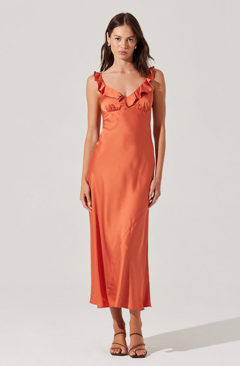 A woman is wearing a sleeveless, floor-length satin dress in a warm orange color. The dress features ruffled straps and a V-neckline, with a fitted bodice that flows into a slightly flared hem. She is also wearing strappy high-heeled sandals.