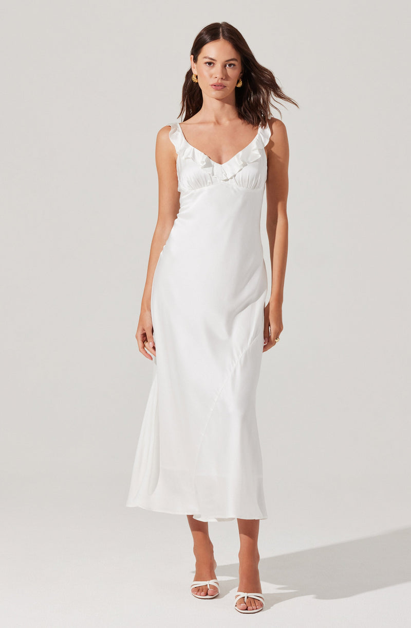 The dress is a sleeveless, full-length gown in white with a V-neckline. It features ruffled detailing along the neckline and straps. The fabric appears smooth and slightly shiny, giving it an elegant, satin-like look. The hemline reaches the ankles, and the dress has a fitted silhouette that slightly flares at the bottom. The woman is wearing open-toe white heels with a thin strap across the toes.