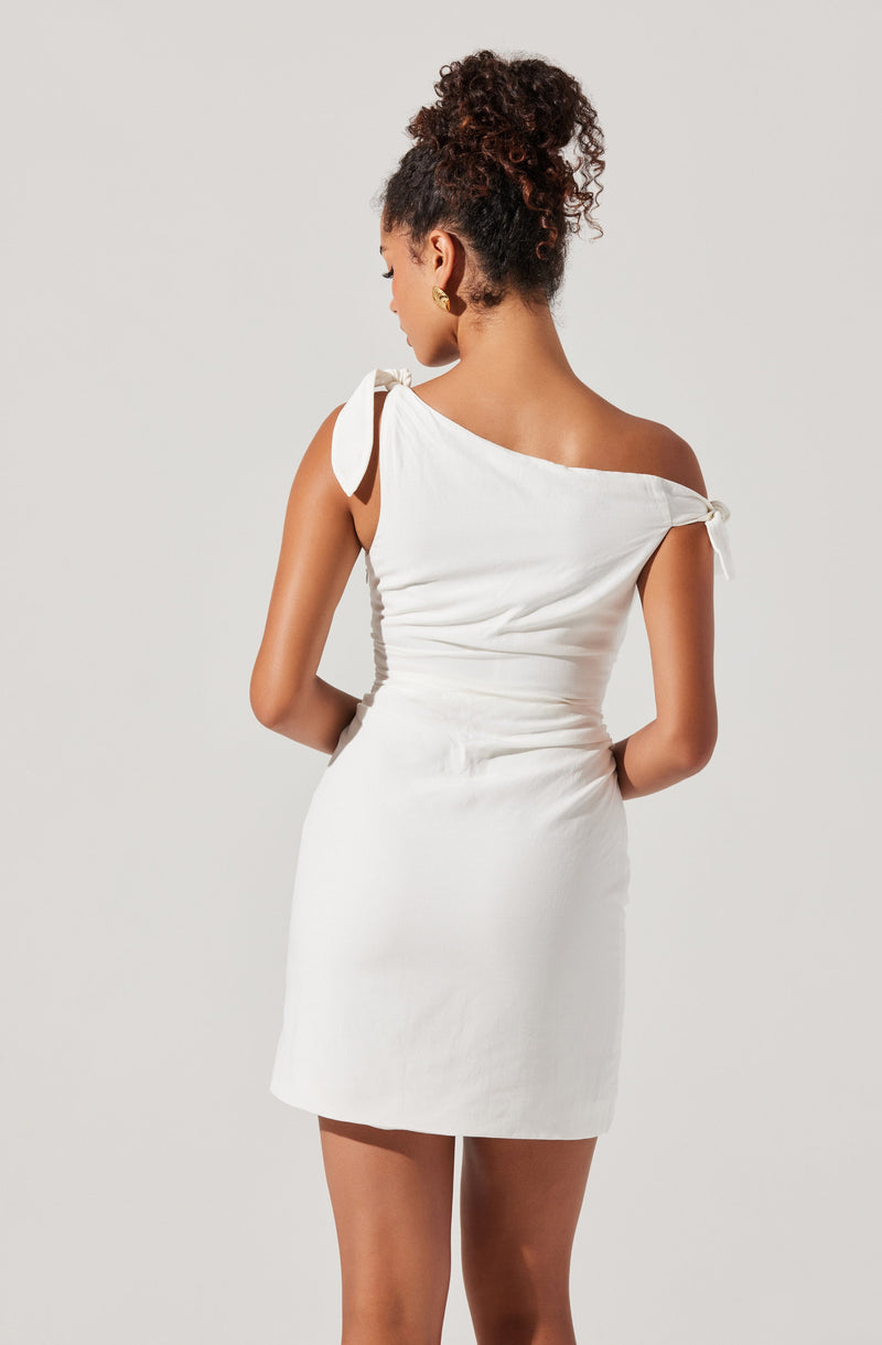 A woman is wearing a white, sleeveless, mini dress with one shoulder strap tied into a bow and the other shoulder strap draped off the shoulder. The dress appears to be made of a satin-like material and has a fitted silhouette. She is also wearing gold hoop earrings and has curly hair styled in an updo.