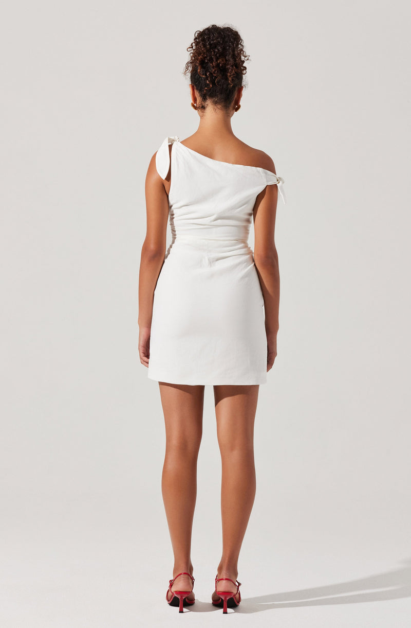 A woman is wearing a short, white, one-shoulder dress with a decorative bow on the shoulder strap. The dress is fitted at the waist and has a simple, straight skirt. She is also wearing red high-heeled shoes.