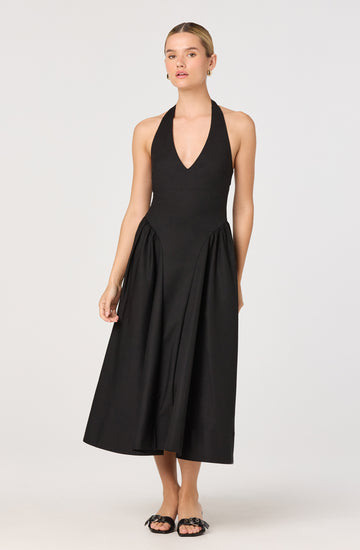 A woman is wearing a sleeveless black halter neck dress with a deep V neckline. The dress has a fitted bodice and a full, gathered skirt that falls to mid-calf length. She is also wearing black open-toe sandals with decorative embellishments.