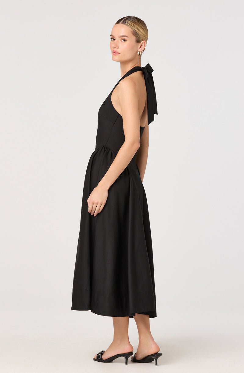 A woman is wearing a sleeveless black halter neck dress with a bow tied at the back of the neck. The dress has a fitted bodice and a flowy, ankle-length skirt. She is also wearing black open-toe heels with a slight heel height.