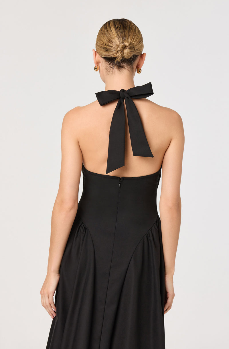 A woman is wearing a strapless black gown with a fitted bodice and a full skirt. The dress has a zipper closure at the back. She has a large black bow tied around her neck, with the ribbons hanging down her back. She also has gold hoop earrings and her blonde hair is styled in a sleek, low bun.