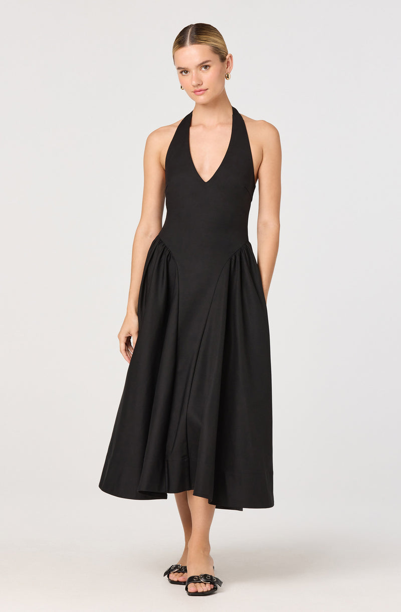 A woman is wearing a sleeveless, black halter dress with a deep v-neckline. The dress has a fitted bodice and a full, gathered skirt that falls to mid-calf length. She is also wearing black open-toe sandals with decorative embellishments on the straps.