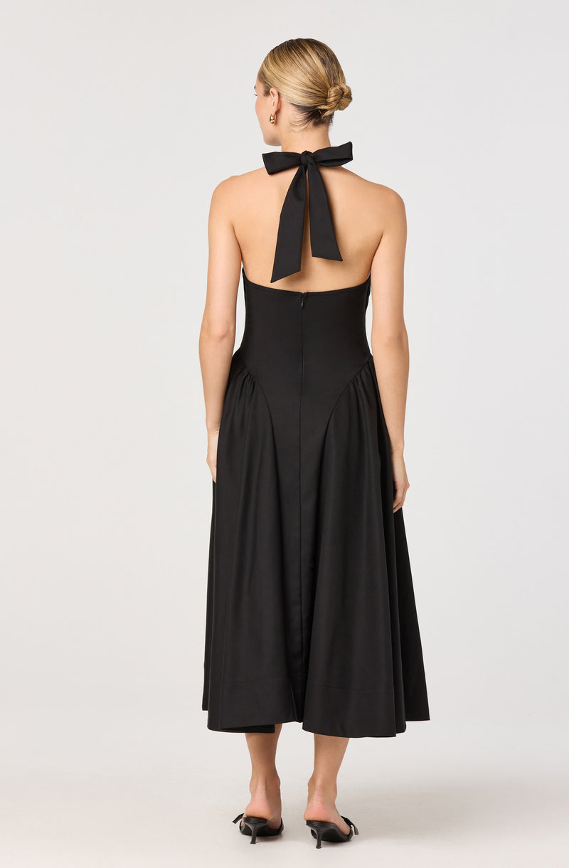 The woman is wearing a strapless black dress with a fitted bodice that extends to a full, pleated, midi-length skirt. The dress features a large bow tied around the back of her neck with long tails draping down her back. She is also wearing black high-heeled shoes.
