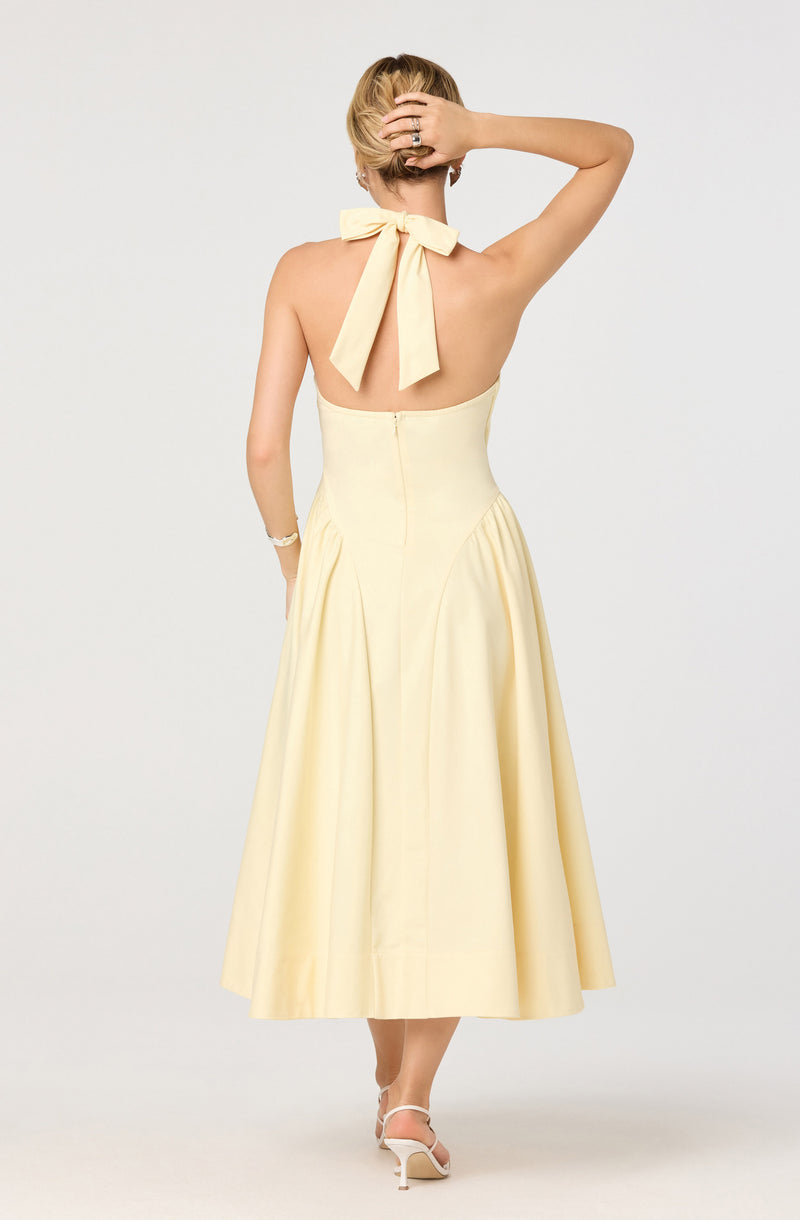 A woman is wearing a strapless, light yellow dress with a fitted bodice that extends to a full, mid-calf length skirt. She has a large, matching yellow bow tied around her neck, with the ends hanging down her back. She is wearing white high-heeled shoes and a bracelet on her right wrist. Her short, blond hair is styled casually, and she is posed with one hand resting on her head and the other hanging by her side.