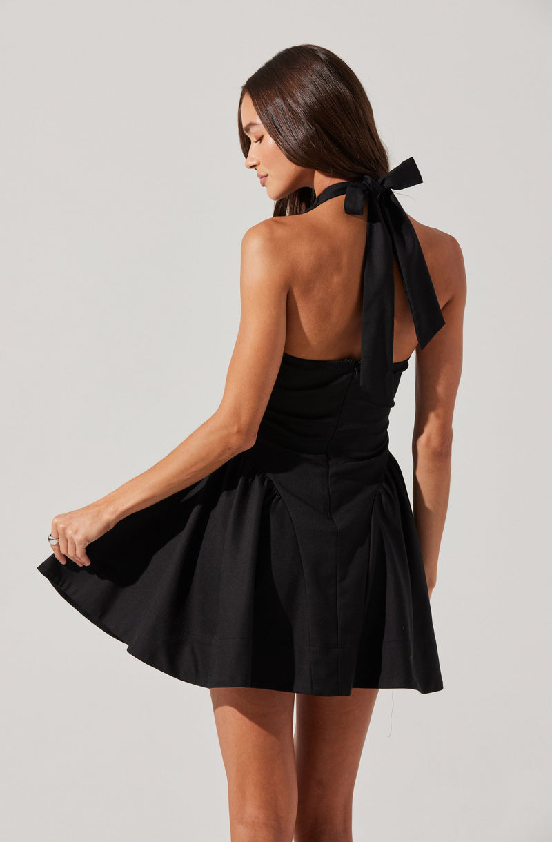 A woman is wearing a black strapless dress with a fitted bodice and a full, flared skirt. The dress features a halter neckline with a large black bow tied at the back of her neck. The fabric of the dress has a slight sheen and is pleated around the waist, creating a voluminous silhouette.