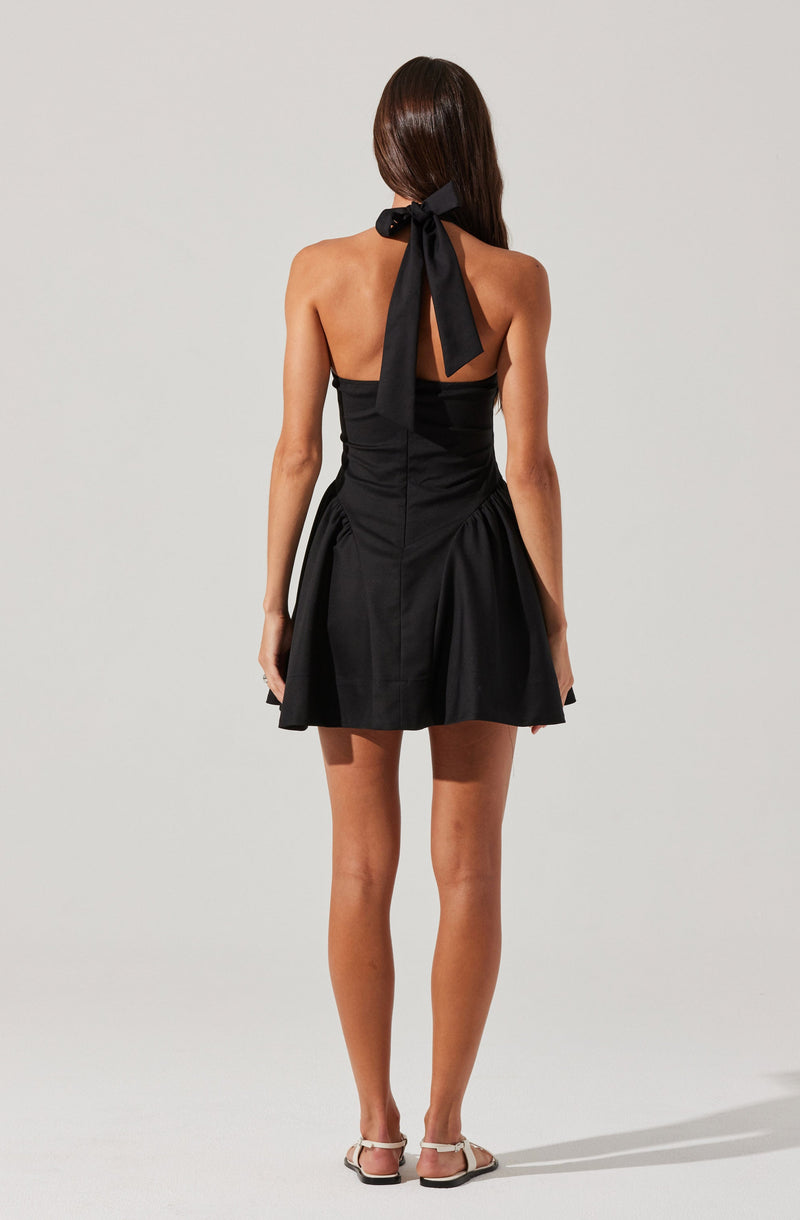 A woman is wearing a black strapless dress with a fitted bodice and a flared skirt that has gathered details at the sides. The dress features a large black ribbon tied into a bow at the back of the neck, and she is wearing flat, open-toed sandals with ankle straps.
