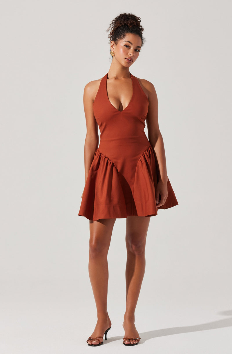 A woman is wearing a sleeveless, rust-colored dress with a fitted bodice and a deep V neckline. The dress has a gathered, flared skirt with pleats that add volume and movement. She is also wearing a pair of black, strappy high-heeled sandals.