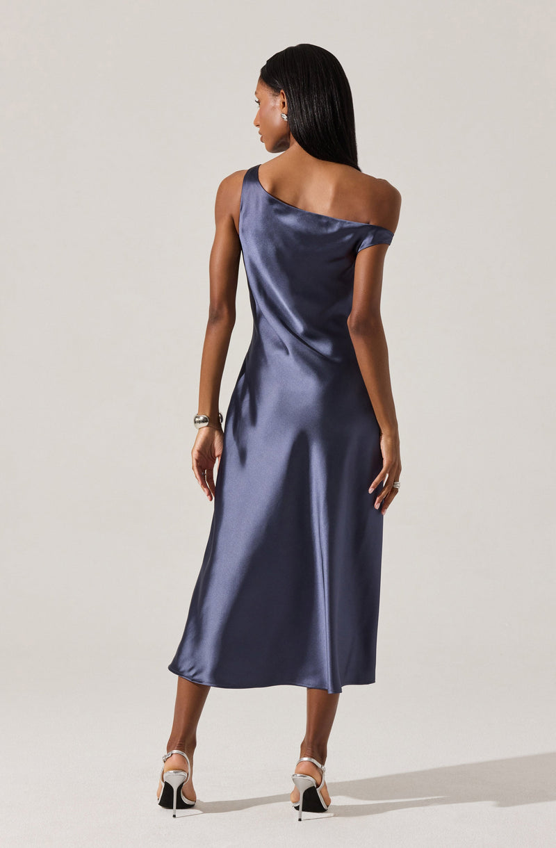 A woman is wearing a floor-length, satin dress in a deep navy blue color. The dress features a one-shoulder design with a draped, off-the-shoulder sleeve on one side and a sleeveless strap on the other. The fabric has a shiny, smooth appearance with a slight sheen, and it falls elegantly to her ankles. She is accessorized with a bracelet on her left wrist and a ring on her right hand, and she is wearing high-heeled shoes with ankle straps.