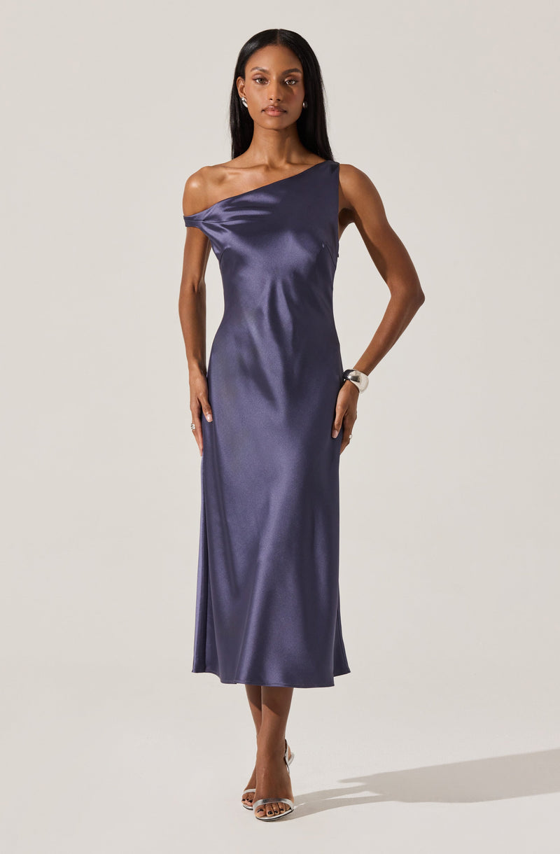 A woman is wearing a dark blue, satin, one-shoulder gown that drapes over her body, reaching down to her ankles. The dress features a loose, elegant fit with a single off-the-shoulder strap on the left side. She is accessorized with earrings, a watch, and open-toe heels.