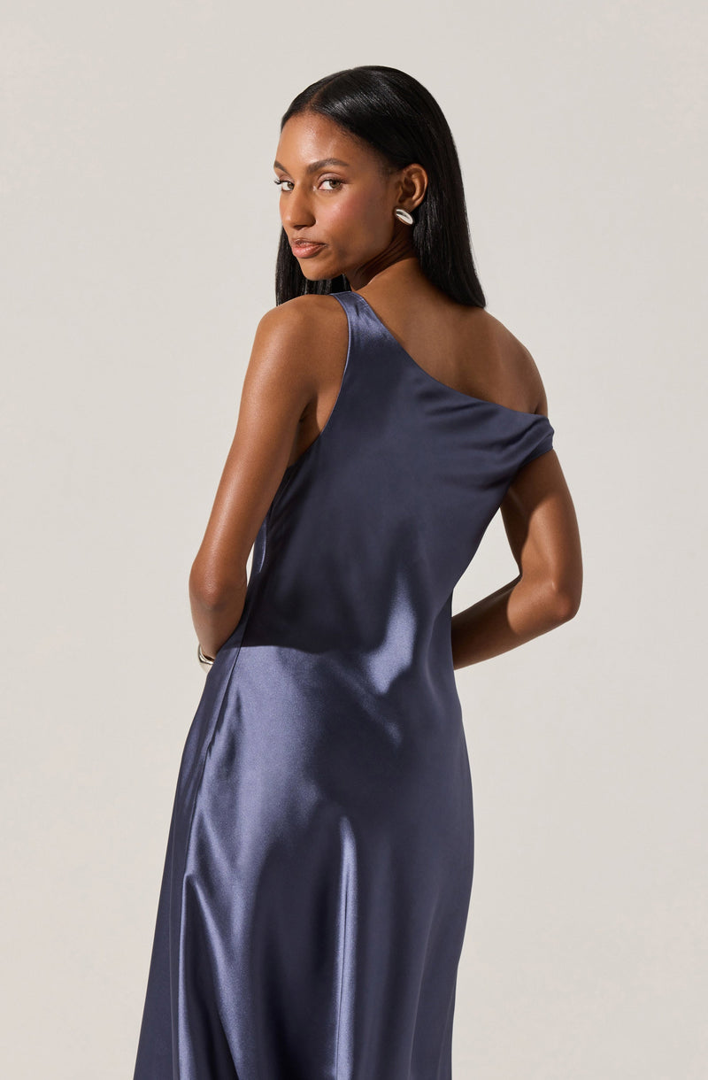 A woman wearing a sleeveless, silky navy blue slip dress with a wide off-the-shoulder neckline, fitted at the waist and flowing down, paired with silver earrings.