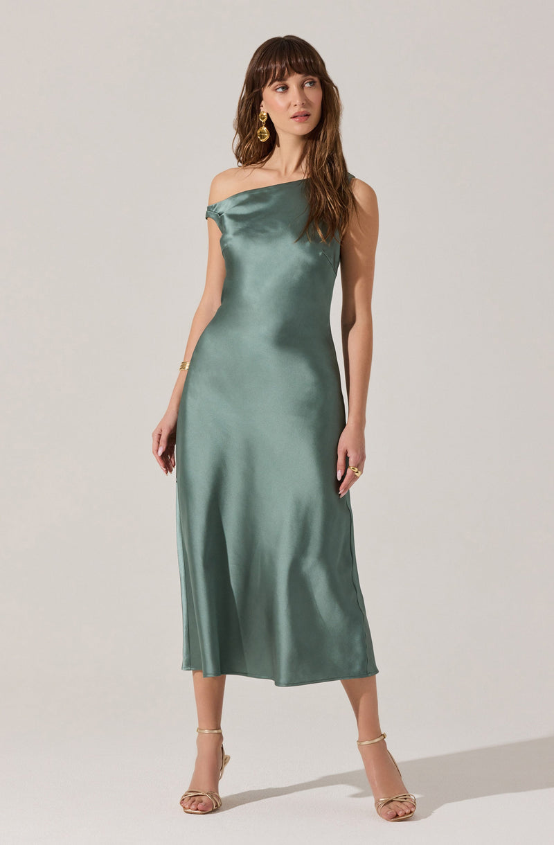 A woman is wearing a silky, satin-like, emerald green, off-the-shoulder dress that falls to her mid-calf. The dress has a smooth, shiny texture and a relaxed fit. She accessorizes with large, gold, hoop earrings, a gold bracelet, and rings, and she is wearing beige, strappy high-heeled sandals.
