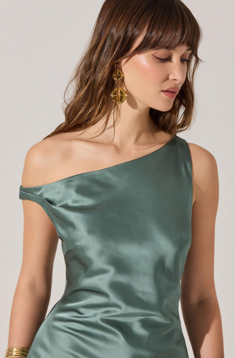 A woman wearing a satin, one-shoulder dress in a muted green color. The dress has a smooth, shiny texture and drapes elegantly across her body, with the right shoulder exposed and the left shoulder covered. She accessorizes with large, gold statement earrings and a gold bracelet.