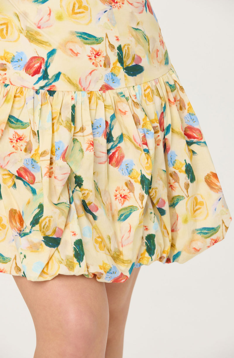 A close-up of a dress with a floral pattern featuring various colors, including yellow, red, blue, green, and pink. The fabric is light and appears to be soft, with gathering at the waist creating a voluminous, bubble-style skirt. The dress has a smooth, rounded waistband and the skirt is slightly puffed at the bottom.